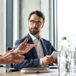serious male ceo listen to female colleague holding notebook at table meeting.