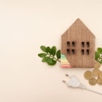 wooden house model with green leaves and coins and power plug on neutral background,sustainable energy and electricity home prices concept
