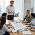 Architektenteam in kurzer Abstimmung an einem Besprechungstisch. Eine Person erklärt aktiv anhand von Unterlagen oder Tablet, während andere aufmerksam zuhören.