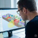 Architekt sitzt leicht nach vorne geneigt und analysiert ein Modell am Bildschirm. Die Darstellung zeigt eine typische Prüf- bzw. Analyseansicht mit farblich differenzierten Bauteilen oder hervorgehobenen Bereichen