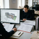 Ein Architekt sitzt konzentriert am Schreibtisch und arbeitet gleichzeitig mit einem Bildschirm, auf dem eine Archicad-nahe Modellansicht sichtbar ist, sowie mit ausgedruckten Tabellen, Notizen und strukturierten Projektunterlagen.